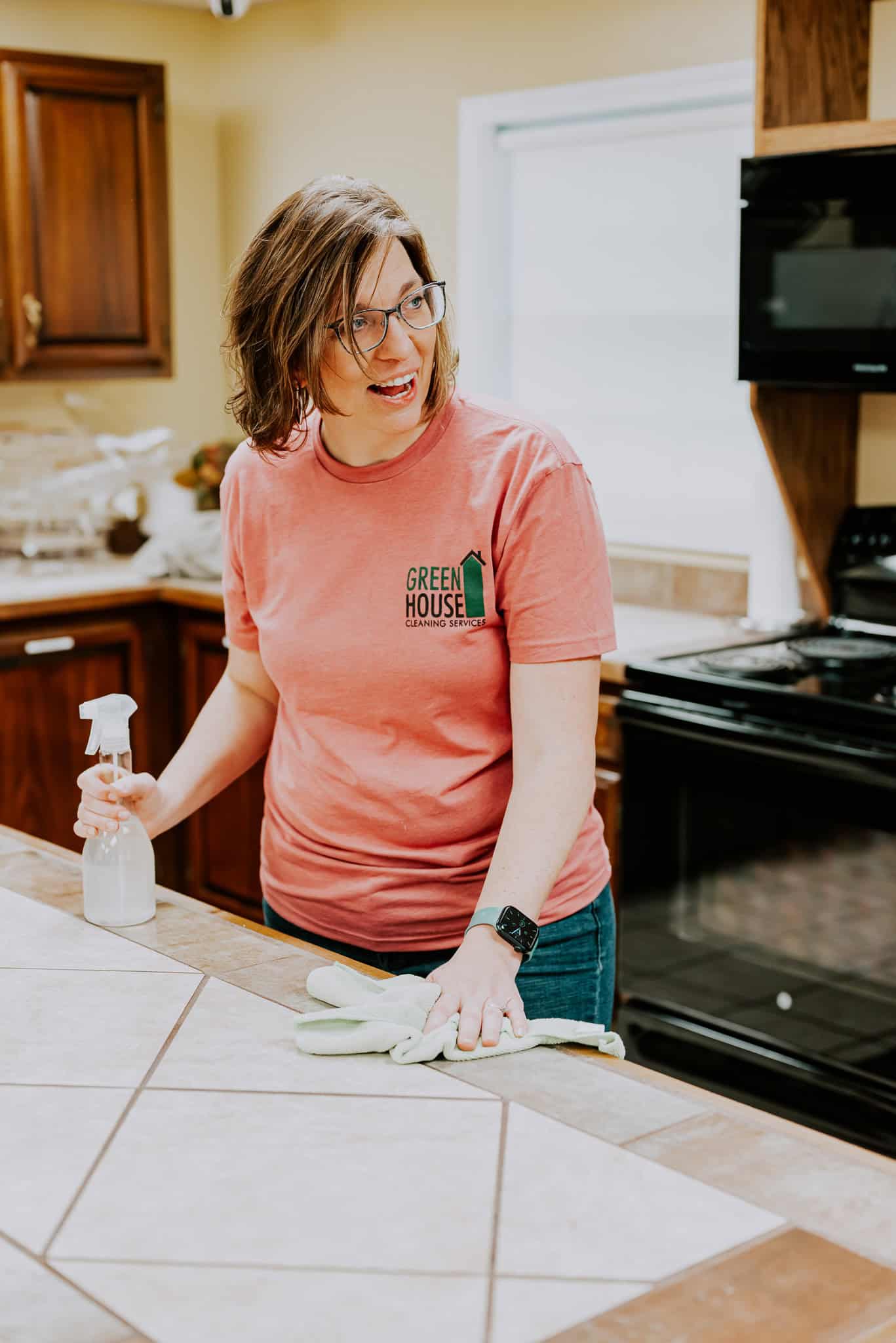 Kim cleaning a kitchen counter in a Green House Cleaning shirt